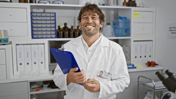 Un hombre sonriente con una barba de pie en un laboratorio con una bata de laboratorio y sosteniendo un portapapeles. - Foto, Imagen
