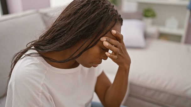 Stresli, kıvırcık saçlı Afro-Amerikan bir kadın evde oturuyor ve endişe dolu duygular gösteriyor.. - Fotoğraf, Görsel