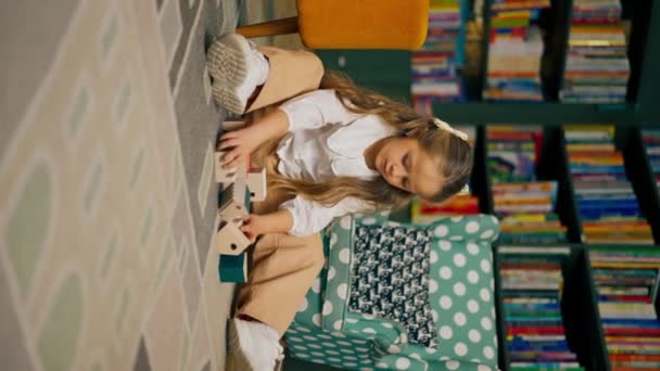 pionowe wideo zbliżenie w księgarni w strefie dla dzieci piękna długowłosa dziewczyna bawi się drewnianymi zabawkami - Materiał filmowy, wideo