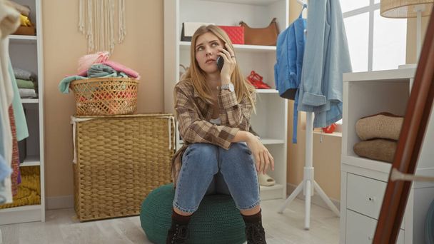 Una giovane donna contemplativa parla su uno smartphone seduta in uno spogliatoio elegante e disorganizzato. - Foto, immagini