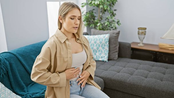 Besorgte junge blonde Frau mit Bauchschmerzen, auf dem Wohnzimmersofa sitzend, zu Hause ausgeruht, ein Porträt ihres schlechten Zustandes im Haus - Foto, Bild