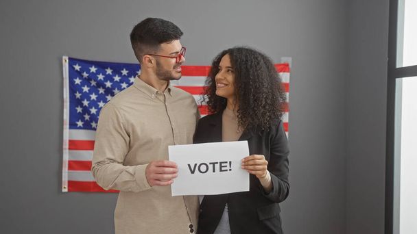 Мужчина и женщина в профессиональной одежде стоят в помещении с флагом США, держа табличку с надписью "Голосуйте!" - Фото, изображение