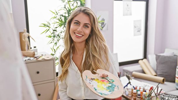 Una giovane artista bionda sicura di sé, irradiata di gioia, brandisce una tavolozza in uno studio d'arte al coperto mentre si immerge nella creazione di dipinti e nella gioiosa creatività - Foto, immagini