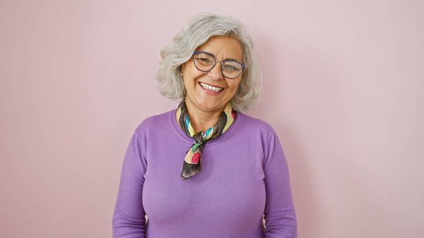 Una mujer madura alegre con el pelo gris, gafas, y con un suéter púrpura, posa sobre un fondo rosa - Foto, imagen