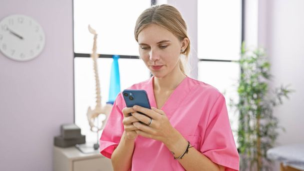 Blondýny žena v růžové pračky pomocí smartphonu v místnosti kliniky s lékařským vybavením v pozadí. - Fotografie, Obrázek