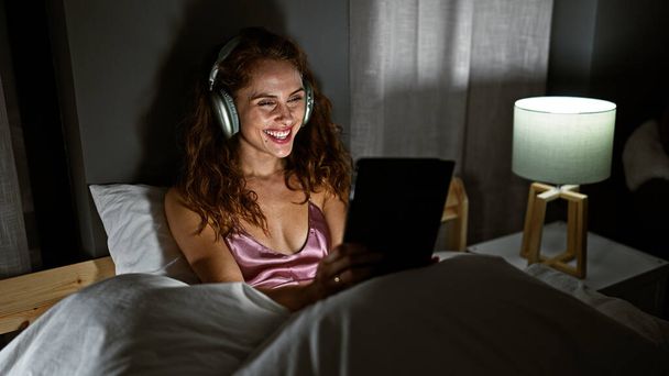 A smiling young woman enjoys her tablet in bed at night while wearing headphones, presenting a cozy, domestic scene. - Photo, Image