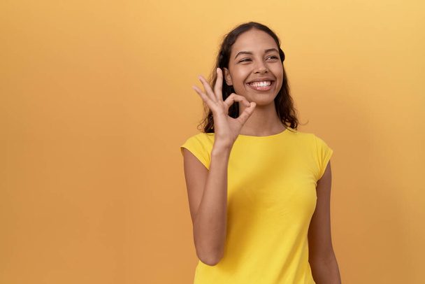 Joven africana americana sonriendo confiada haciendo ok signo sobre aislado amarillo fondo - Foto, Imagen