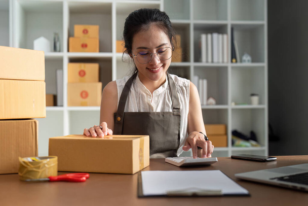 A woman is sitting at a desk with a calculator and a stack of boxes. She is smiling and she is happy - Photo, Image