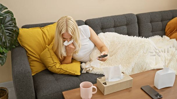 Oszałamiająca młoda blondynka, chora na paskudne przeziębienie, leży pod przytulnym kocem na swojej domowej sofie, kichając i karmiąc filiżankę ciepłej herbaty podczas oglądania swoich ulubionych programów telewizyjnych. - Zdjęcie, obraz