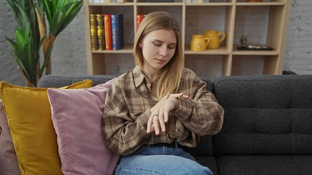 Egy fiatal nő ellenőrzi az időt, miközben ül egy kanapén egy hangulatos nappaliban, felidézve egy nyugodt, otthonos hangulatot. - Fotó, kép