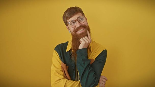 Un uomo con la barba rossa sorridente in casualwear posa pensieroso su uno sfondo giallo. - Foto, immagini