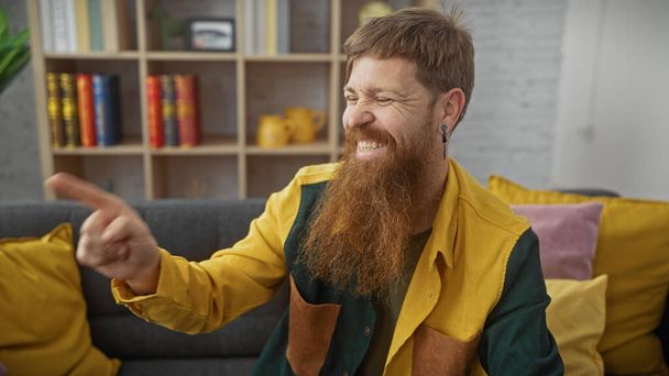 Rire rousse barbu homme pointant du doigt tandis qu'il était assis sur un canapé jaune à l'intérieur. - Photo, image