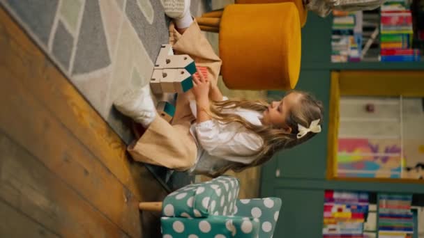 子供エリアの本屋で垂直ビデオクローズアップ 美しい長髪の少女は木のおもちゃで遊ぶ - 映像、動画