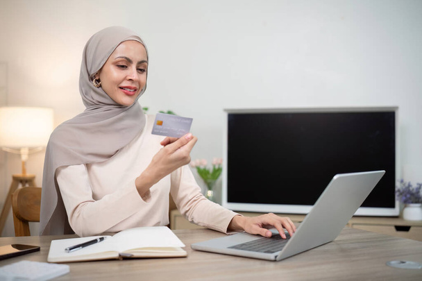 Nuori onnellinen musliminainen hijabissa kotona kannettavan tietokoneen ostoksia verkossa luottokortilla istuessaan pöydällä. - Valokuva, kuva