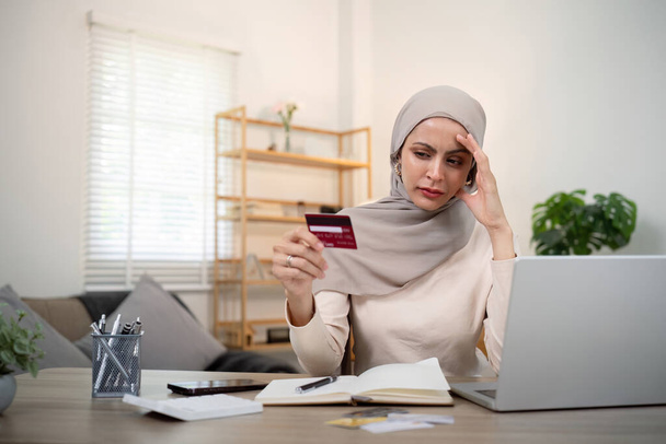 Πρόβλημα πιστωτικής κάρτας. Ανησυχία και άγχος νεαρή μουσουλμάνα γυναίκα σε hijab κάθεται στο σαλόνι εργασίας και να κάνουν την αγορά στο ηλεκτρονικό κατάστημα για laptop. - Φωτογραφία, εικόνα