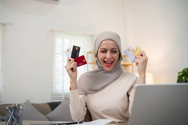Νεαρή ευτυχισμένη μουσουλμάνα γυναίκα σε hijab στο σπίτι χρησιμοποιώντας φορητό υπολογιστή ψώνια σε απευθείας σύνδεση με πιστωτική κάρτα, ενώ κάθεται στο γραφείο. - Φωτογραφία, εικόνα