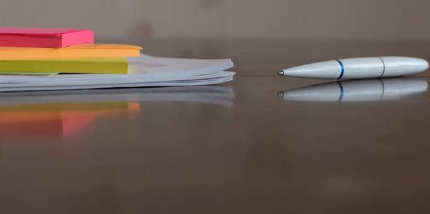 Разноцветные бумажные наклейки и белая шариковая ручка крупным планом на деревянном столе с отражениями на размытом фоне - Фото, изображение