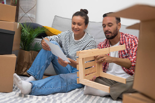 Gli sposi felici installano mobili in soggiorno che si siede su pavimento tra decorazioni raccolte su territorio di appartamento moderno ricevuto come regalo nuziale da genitori - Foto, immagini