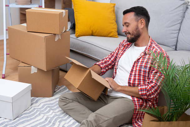 New owner with smile holds carton box in hands sitting on floor with covered surface emitting cheerful emotions on face rejoicing in purchase of modern apartment - Photo, Image