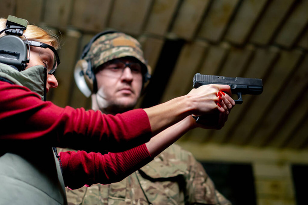 χαμηλή βολή σε ένα επαγγελματικό σκοπευτήριο ένας στρατιωτικός λέει και δείχνει σε ένα κορίτσι τη σωστή στάση με ένα πιστόλι είναι - Φωτογραφία, εικόνα