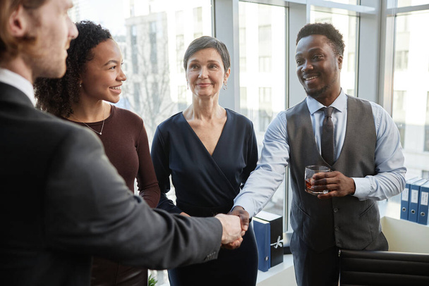 Diverso grupo de socios de negocios sonrientes estrechando la mano en la oficina de pie con bebidas y celebrando trato - Foto, imagen