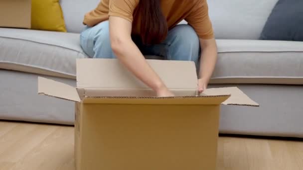 Donna esamina con interesse vestito colorato da scatola di cartone dal negozio online con abbigliamento femminile gioisce al pacco mentre seduto sul divano in appartamento - Filmati, video