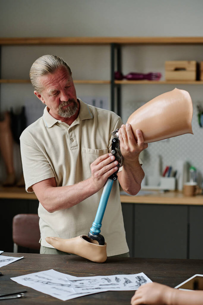 Retrato vertical do artesão sênior de cabelos compridos construindo próteses de perna na oficina e inspecionando articulações - Foto, Imagem