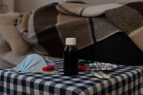 Eldobható orvosi maszk, hőmérő, bájitalok és tabletták homályos háttér a személy alatt a kockás kanapén - Fotó, kép