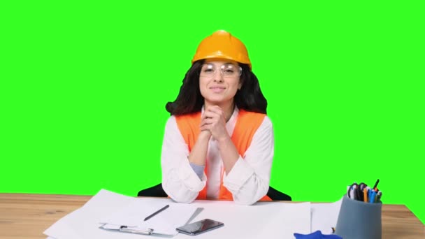 Smiling young female engineer in protective helmet working with drawing, looking at camera on green isolated background. Industry, engineering concept - Footage, Video