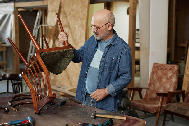 Портрет старшего человека в мастерской по реставрации мебели, фиксирующий старый деревянный стул, копировальное пространство - Фото, изображение