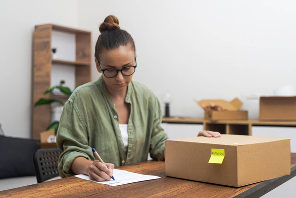 Capturada de cerca, una mujer sentada en su mesa, profundamente centrada en escribir una solicitud de devolución, compras en línea devolver un producto.  - Foto, imagen