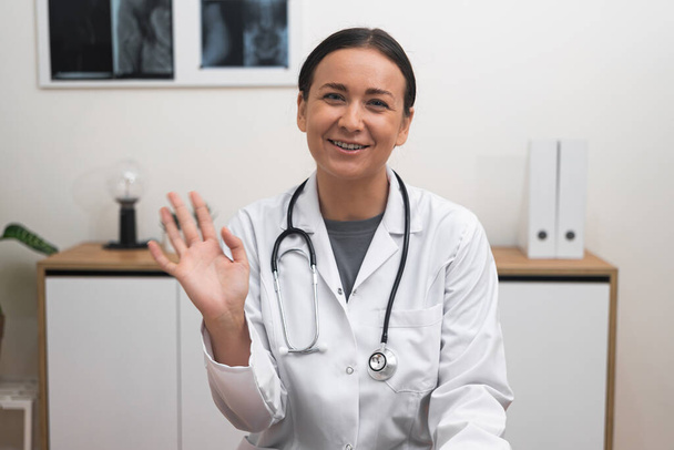 Online Consultatie Welkom: Met een welkome glimlach, een vrouwelijke arts zwaaiende hand houdt zich bezig met de camera, waardoor patiënten zich comfortabel voelen tijdens hun online afspraak.  - Foto, afbeelding