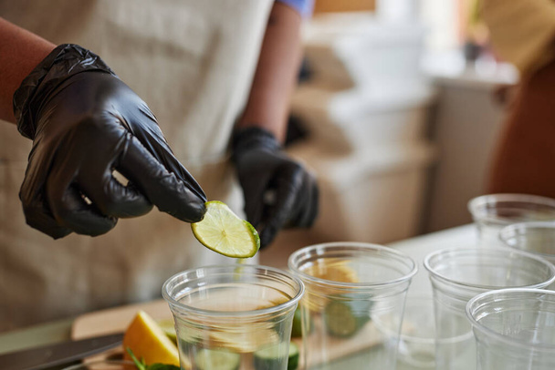 Blisko kobiety ubranej w rękawiczki dodającej plasterki limonki do orzeźwiających napojów lemoniady w barze, przestrzeń do kopiowania - Zdjęcie, obraz