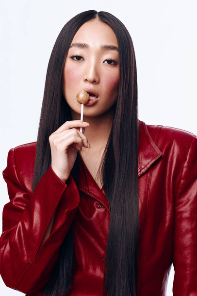 Elegante donna asiatica in giacca di pelle rossa con lecca-lecca in bocca in ambiente urbano - Foto, immagini