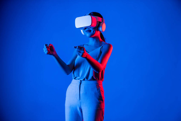 Inteligentna damska podstawka otoczona światłem neonowym noszącym zestaw słuchawkowy VR łączący metawertyczną, przyszłą technologię społeczności cyberprzestrzeni. Elegancka kobieta cieszyć się gry wyścigowe w meta świecie. omamy. - Zdjęcie, obraz