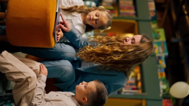 vídeo vertical em uma livraria na área de crianças menina bonita e menino sua mãe para lê-los um livro - Filmagem, Vídeo