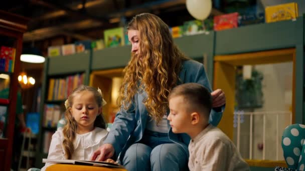 в книжном магазине в детской зоне красивые девочка и мальчик их мать, чтобы прочитать им книгу - Кадры, видео