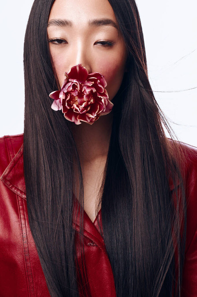 Mujer misteriosa con el pelo largo y negro y chaqueta de cuero rojo sosteniendo una flor delante de su cara - Foto, Imagen