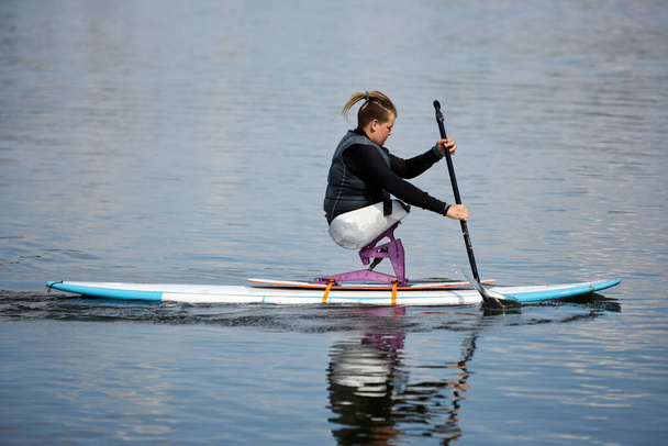 Visão lateral retrato de jovem adolescente com deficiência apreciando paddleboarding no lago com equipamento esportivo adaptativo, espaço de cópia - Foto, Imagem