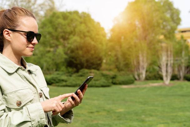 Donna in occhiali da sole messaggistica online utilizzando il suo smartphone mentre cammina parco cittadino in primavera. - Foto, immagini