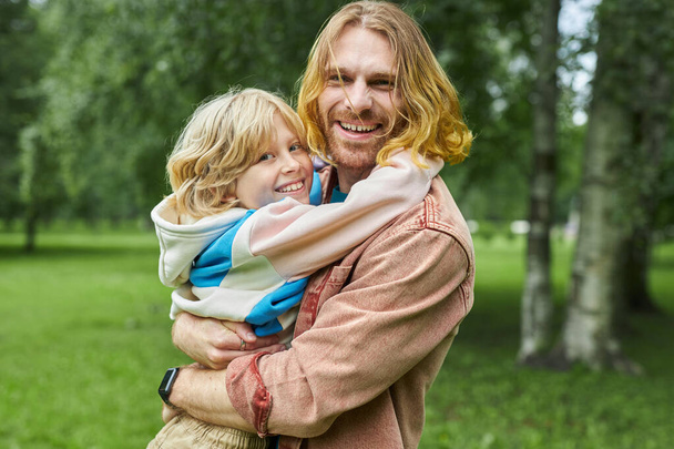 Porträt des glücklichen Vaters, der einen blonden kleinen Jungen im Park hält und gemeinsam in die Kamera schaut - Foto, Bild