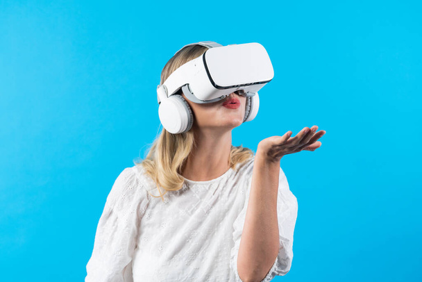 Blank meisje met VR-bril en staand terwijl ze iets vasthoudt en op een blauwe achtergrond staat. Vrouw blazend en kijkend naar hologram in handen terwijl ze de visuele werkelijkheid verbindt. Contraptie. - Foto, afbeelding