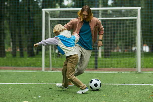 Plan d'action d'un garçon blond jouant au football avec son père à l'extérieur et donnant des coups de pied, espace de copie - Photo, image