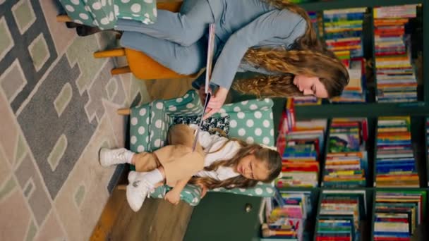 függőleges videó egy könyvesboltban a gyerekek területén gyönyörű lány és fiú nem akarja, hogy az anyjuk olvasson nekik egy könyvet - Felvétel, videó