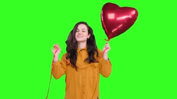 Usmívající se mladá žena pózuje se vzduchovým balónem ve tvaru srdce a tančí na zeleném izolovaném pozadí. Milostný nápad. Valentýn - Záběry, video