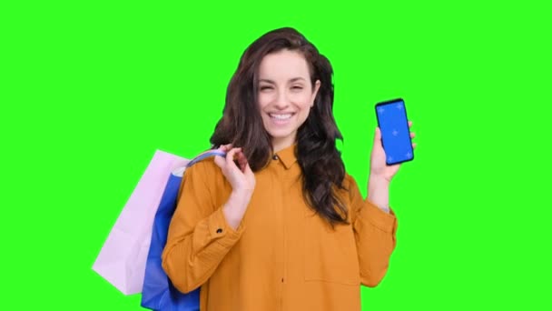 Mujer joven y feliz posando con bolsas después de comprar, mostrando la pantalla del teléfono móvil en un fondo verde aislado. Concepto de estilo de vida - Imágenes, Vídeo