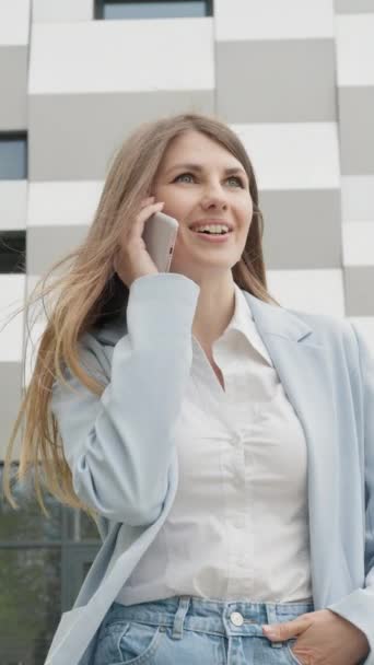 Вертикальний екран: Кавказька впевнена молода ділова жінка в білій сорочці розмовляє по телефону зовні біля сучасної будівлі офісу. Середній довгий низький кут 4K Повільний рух Корпоративний знімок з переміщенням навколо - Кадри, відео