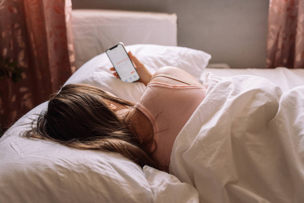 La chica se siente mal y se acuesta en la cama sosteniendo un teléfono con un calendario en la mano. - Foto, imagen