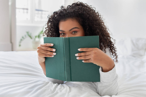 Una joven hispana con el pelo rizado está parcialmente escondida detrás de un libro verde mientras lee, acostada boca abajo sobre una cama blanca con una mirada de intriga o diversión - Foto, imagen