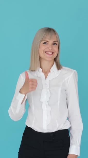 Cheerful Caucasian woman showing thumbs up hand sign, standing on blue background. Good choice, positive gesture concept. Vertical video. - Footage, Video